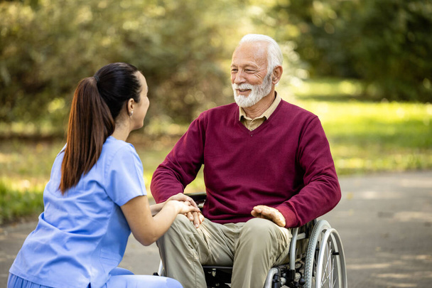 Θηλυκή νοσοκόμα με στολή που φροντίζει ηλικιωμένο άνδρα σε αναπηρική καρέκλα. - Φωτογραφία, εικόνα