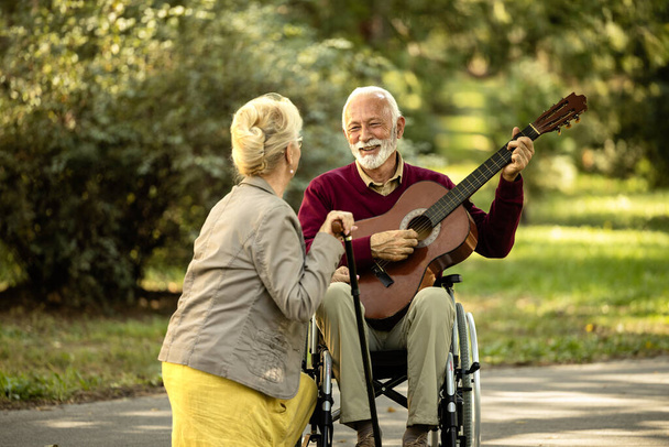 Ηλικιωμένος άνδρας σε αναπηρική καρέκλα παίζει κιθάρα και απολαμβάνει τη στιγμή με τη σύζυγό του. - Φωτογραφία, εικόνα