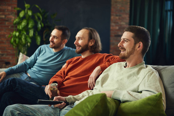 Ευτυχισμένοι και χαρούμενοι νέοι άνδρες με τηλεχειριστήριο και βλέποντας ποδοσφαιρικό αγώνα καθισμένοι σε πούλμαν στο σαλόνι στο σπίτι. Έννοια της φιλίας, ομαδικότητα, τηλεοπτικό πρόγραμμα, online εκδήλωση, αναψυχή. - Φωτογραφία, εικόνα