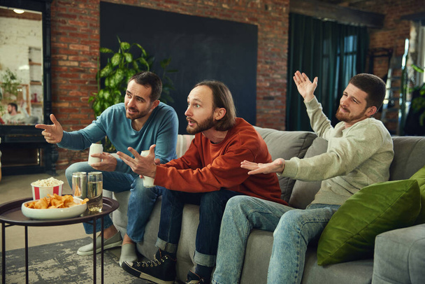 Happy and cheerful young men with alcohol drinks and snacks watching football match sitting on coach in living room at home. Concept of friendship, togetherness, TV program, online event, recreation. - Photo, Image