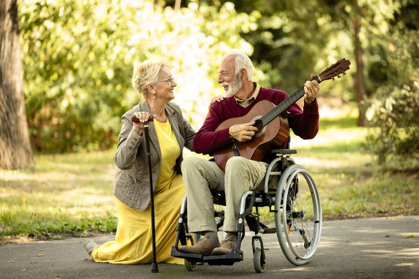 シニアカップルの愛と幸せ. 彼の素敵な妻にギターを弾く車椅子の高齢男性. - 写真・画像