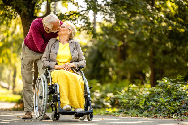 Літня пара закохана. Старший чоловік цілує свою дружину в інвалідному візку в дитячому будинку-парку. - Фото, зображення
