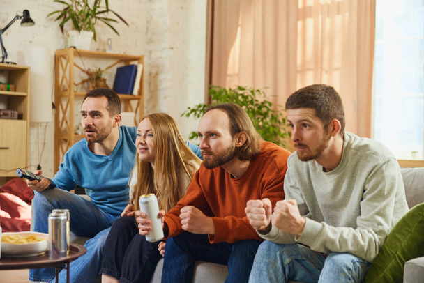 Attractive young people, man and woman sitting in front of TV and attentively watching show, football match and eating snacks with alcohol drinks. Concept of friendship, togetherness, online event. - Photo, Image