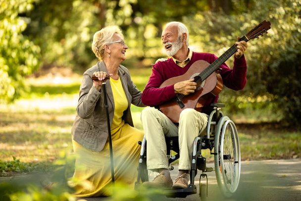 Miłość i szczęście pary seniorów. Starszy mężczyzna na wózku inwalidzkim gra na gitarze do swojej uroczej żony. - Zdjęcie, obraz
