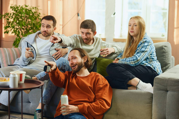 Foto van vrolijke jonge mensen, vrienden die in de woonkamer zitten en zorgvuldig comedy film kijken en praten over acteren. Concept van vriendschap, saamhorigheid, TV-programma, online evenement, recreatie. - Foto, afbeelding