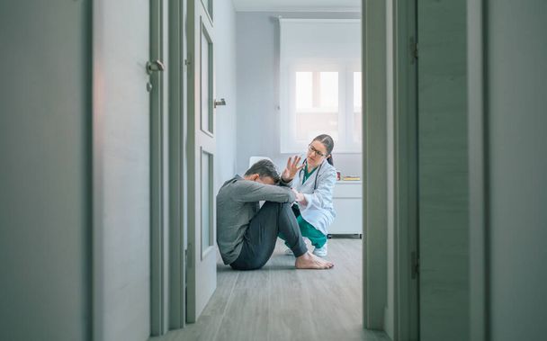 Γυναίκα γιατρός προσπαθεί να βοηθήσει αρσενικό ασθενή με ψυχική διαταραχή που αρνείται να βοηθήσει κάθεται στο πάτωμα του δωματίου της ψυχιατρικής - Φωτογραφία, εικόνα