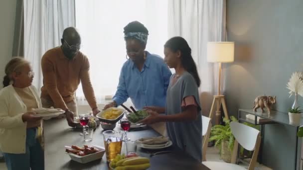Padres afroamericanos felices y dos hijas sirviendo platos en la mesa mientras se preparan para la celebración familiar en casa - Metraje, vídeo