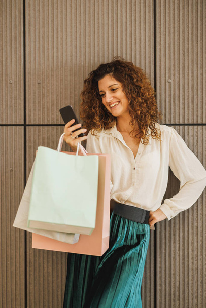 Φωτογραφία μιας γυναίκας που κρατάει τσάντες με ψώνια ενώ χρησιμοποιεί το smartphone της σε φόντο τοίχου σε εμπορικό κέντρο. - Φωτογραφία, εικόνα