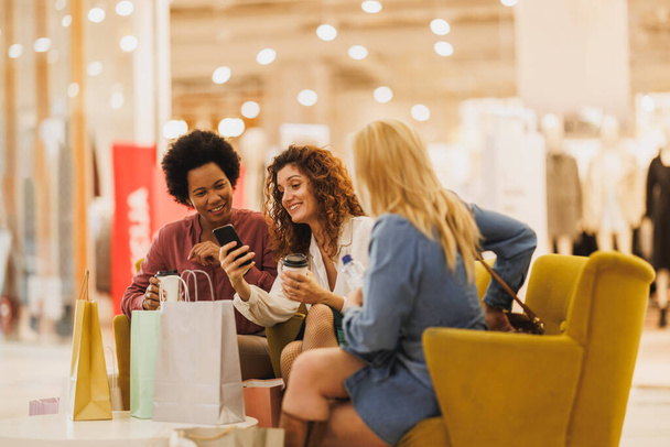 Birlikte alışveriş merkezinde alışveriş yaparken internetten satış tekliflerini kontrol etmek için cep telefonu kullanan üç mutlu kadının fotoğrafı.. - Fotoğraf, Görsel