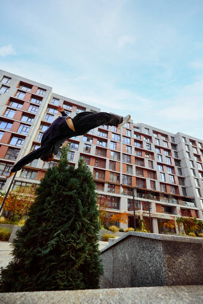 Sportler, junger Mann, der hoch springt, Mauerüberschläge im öffentlichen Park zwischen Hochhäusern über dem Himmel in Aktion. Augenfischfilter. Konzept von Lifestyle, Sport, Freestyle, Aktivität, Bewegung. Anzeige - Foto, Bild