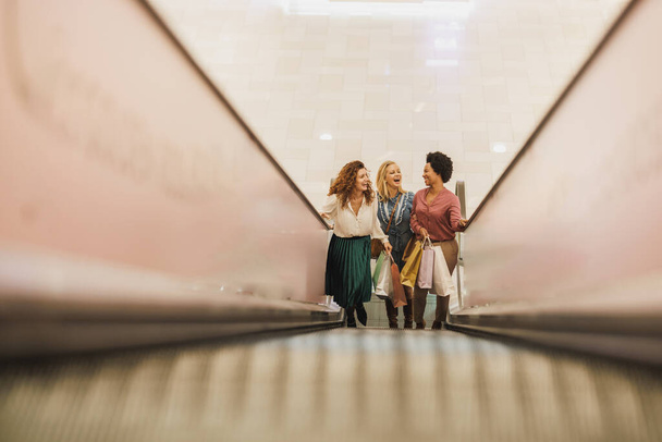 Girato di tre ragazze cheerfu divertirsi e in piedi sulla scala mobile in un centro commerciale della città insieme mentre fuori su una furia dello shopping. - Foto, immagini