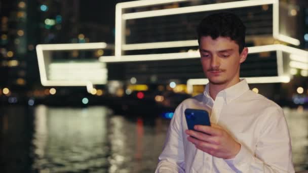 Guy Tourist χρησιμοποιώντας Smartphone, ενώ στέκεται σε Embankment φόντο. Millennial άνθρωπος που ασχολούνται με κύλιση και δακτυλογράφηση στην οθόνη στο δρόμο Night City. Αρσενικό Περιήγηση στο Internet στην κινητή συσκευή - Πλάνα, βίντεο