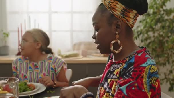 Afroamerikanerin in Kardashiki und Kopftuch beim festlichen Essen und Gespräch mit kleiner Tochter, während sie zu Hause mit der Familie Kwanzaa feiert - Filmmaterial, Video