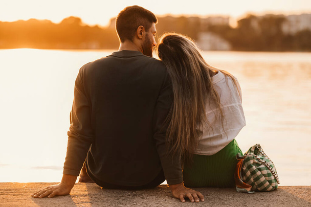 Bel colpo di una bella coppia seduta e godersi il tramonto vicino a un lago della città. - Foto, immagini