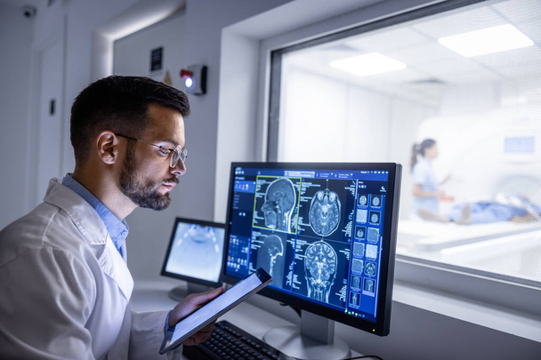 Arzt untersucht Röntgenbilder im MRT-Kontrollraum, während im Hintergrund Krankenschwester den Patienten auf den Untersuchungstest vorbereitet. - Foto, Bild