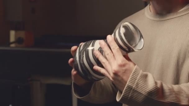 Lassú portré a fiatal kaukázusi férfi üvegfúvóról, ahogy fényképezőgéphez pózol minimalista fekete-fehér mintás üvegvázával, amit maga készített a műhelyben. - Felvétel, videó