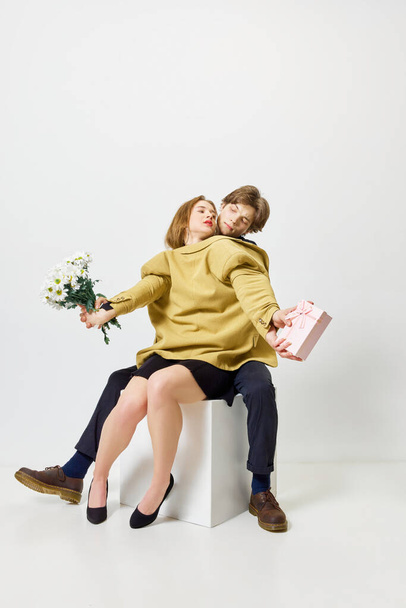 Snoep-boeket periode. Paar knuffels in één jasje met bloemtrossen en aanwezig tegen witte studioachtergrond. Concept van liefde, jeugd, moderne relatie, Valentijnsdag, gevoelens, - Foto, afbeelding
