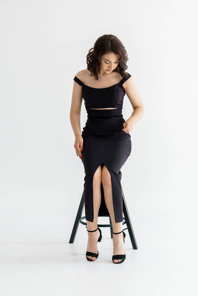 Portrait of a young beautiful girl in a black dress sitting on a chair on a white background - Photo, Image