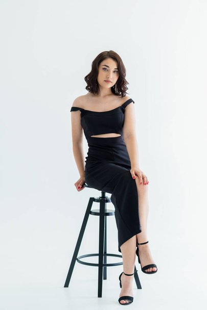 Portrait of a young beautiful girl in a black dress sitting on a chair on a white background - Foto, Bild
