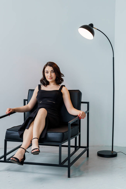 Portrait d'une jeune belle fille en robe noire assise sur une chaise sur fond blanc - Photo, image