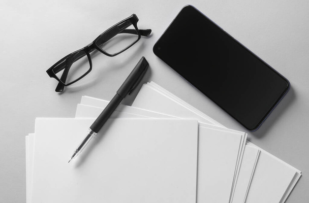 Fogli bianchi vuoti di formato carta a4 o modelli di documenti e smartphone, occhiali e penna su sfondo grigio. Modello per il design. Vista dall'alto. Posa piatta - Foto, immagini