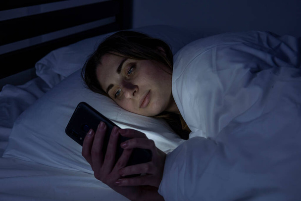 Una ragazza a letto con un telefono su un letto bianco in una stanza buia - Foto, immagini