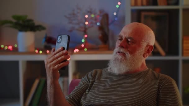 Un hombre de edad avanzada en buen estado de salud y el estado de ánimo utiliza un teléfono inteligente, tiene conversación de videollamada. Una mano de las olas humanas como signo de saludo y felicita a su interlocutor por las vacaciones de Navidad o Año Nuevo - Imágenes, Vídeo