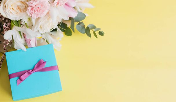Όμορφο μπουκέτο με τριαντάφυλλα, γαρύφαλλα, ίριδα και ευκάλυπτο και μπλε κουτί δώρου με ροζ κορδέλα σε κίτρινο φόντο, αντίγραφο χώρου. Ημέρα μητέρων, 8 Μαρτίου, έννοια δώρο διακοπών - Φωτογραφία, εικόνα