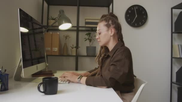 Πλάγια άποψη μέση μέχρι πλάνο της νεαρής καυκάσιας γυναίκας προγραμματιστή με dreadlocks γράφοντας html κώδικα στο ασύρματο πληκτρολόγιο κάθεται μπροστά από την οθόνη του υπολογιστή στο χώρο εργασίας σε μινιμαλιστικό γραφείο - Πλάνα, βίντεο