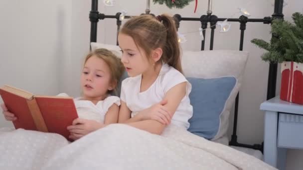 Twee kleine meisjes die een boek lezen op een wit bed, - Video