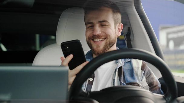Sonriendo joven exitoso sentado en el coche mientras lee el mensaje por teléfono. Estilo de vida, tecnología, concepto de viaje. Movimiento lento - Foto, imagen
