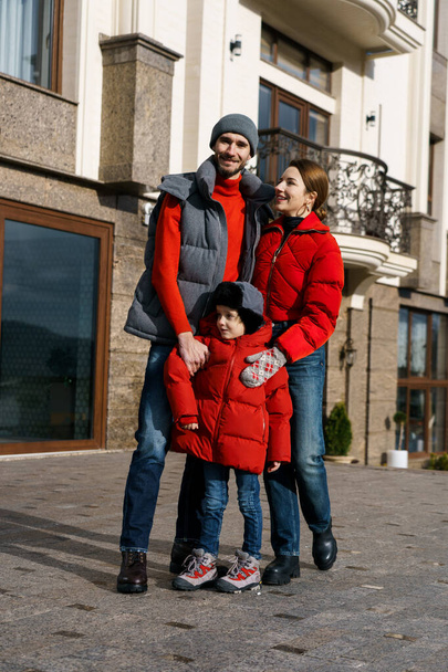 Young family father mother and son posing in red winter clothes near the hotel. Happy family concept - Photo, Image