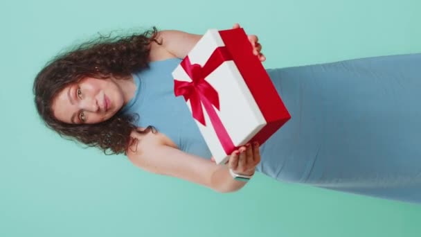 Doğum günü hediyesini sunan, gülümseyen genç bir kadın ellerini uzatıyor, hediye olarak hediye paketi sunuyor, parti satışını kutluyor. Mavi stüdyo arka planında tek başına kalan bir kız. Dikey - Video, Çekim