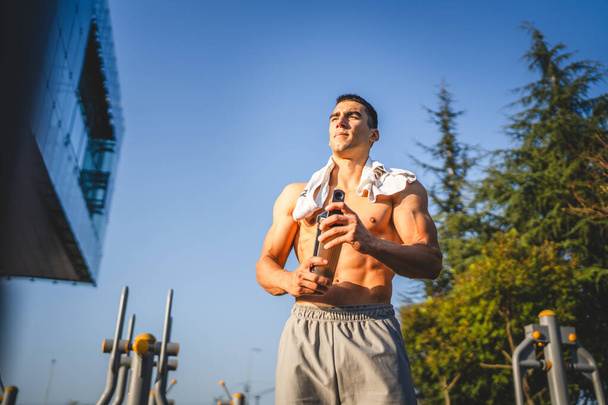 Egy kaukázusi férfi fiatal férfi sportoló, hogy egy fék alatt szabadtéri edzés a parkban szabadtéri tornaterem tartsa kiegészítő shaker a kezében boldog magabiztos erős másolási hely - Fotó, kép