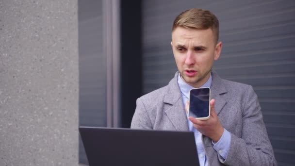 Jeune homme travaillant avec un ordinateur portable tout en parlant sur le smartphone à l'extérieur. Entreprise, emploi, concept technologique. Temps réel - Séquence, vidéo