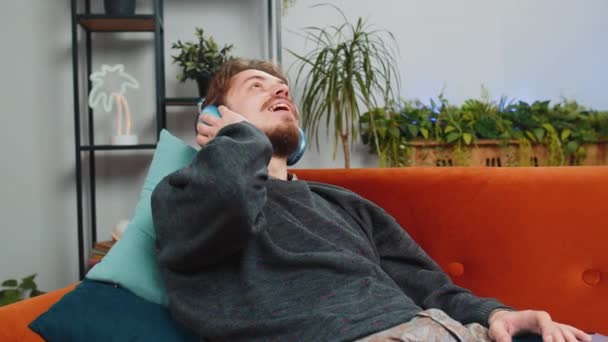 Щасливий розслаблений приємний кавказький молодий чоловік в бездротових навушниках танцює, лежачи на домашньому дивані, вибираючи слухати улюблену енергійну диско-музику в смартфоні. Діяльність вихідних людей - Кадри, відео