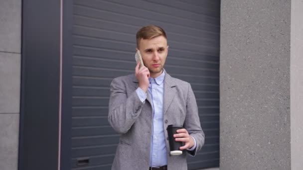 Hymyilevä nuori mies puhuu matkapuhelimella ja juo kahvia, seisoo ulkona. Liiketoiminta, teknologia. Reaaliaikainen - Materiaali, video
