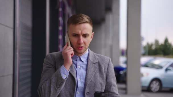 Boze zakenman die koffie vasthoudt terwijl hij buiten loopt en praat op de mobiele telefoon. Zakelijk, werk, technologie concept. Real time - Video