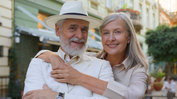 Portrait happy Caucasian family elderly couple husband wife hugging cuddle hug looking at camera outside city street. Retirement senior people enjoy romantic outdoor lifestyle vacations tour together - Photo, Image