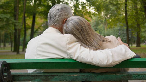 Back view happy lovely gray haired senior married couple sitting bench talk discuss hugging city park outside. Romantic aged man woman embrace talking outdoors enjoying date together retirement relax - Photo, Image