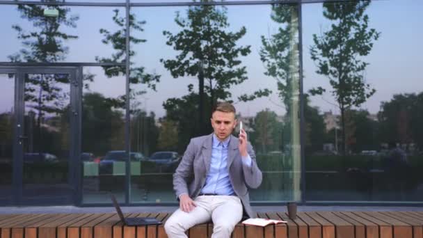 Järkyttynyt liikemies istuu penkillä puhuessaan matkapuhelimeen kaupungissa. Liiketoiminta, työ, teknologia. Reaaliaikainen - Materiaali, video
