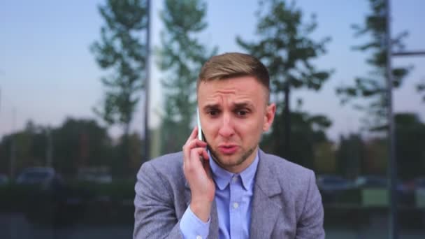 Upset young businessman talking on the smartphone, sitting outdoors. Business, job, technology concept. Real time - Footage, Video