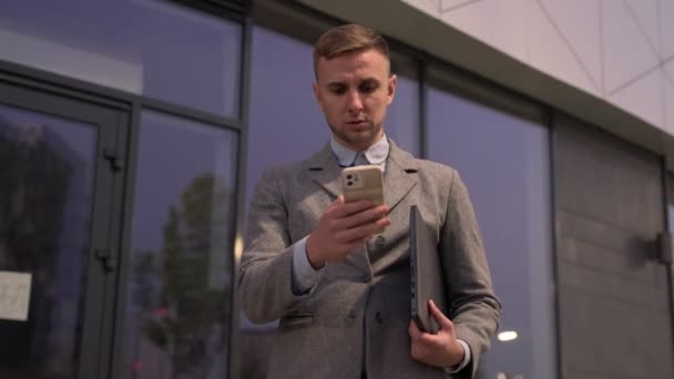 Joven hombre de negocios sosteniendo portátil mientras escribe en el teléfono inteligente, de pie al aire libre. Negocios, concepto tecnológico. Tiempo real - Imágenes, Vídeo
