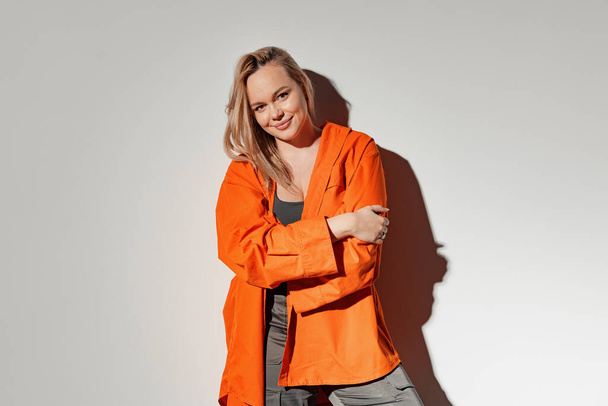 A confident woman in a striking orange jacket poses with a playful smile, exuding style and poise against a light background - Foto, Imagen