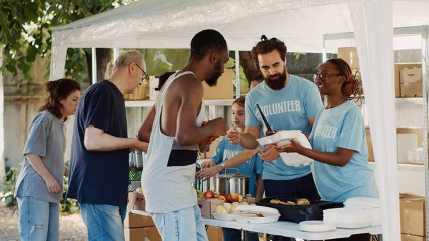 Ομάδα ανθρωπιστικής βοήθειας παρέχει ευγενικά δωρεάν ζεστά γεύματα στους φτωχούς άπορους και λιγότερο τυχερούς ανθρώπους. Ομάδα εθελοντών που μοιράζουν δωρεές τροφίμων στους άστεγους. Σφηνάκι τρίποδα. - Φωτογραφία, εικόνα