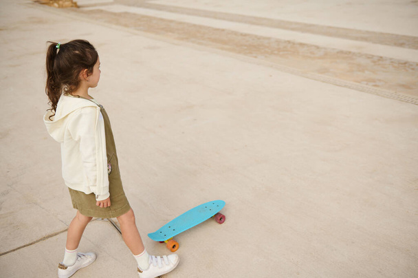 Klein meisje in beige hoodie en kaki jurk, skateboarden op het stedelijke skatepark of speeltuin. Zicht van bovenaf. Gelukkige actieve jeugd. Extreme sport. Vrijetijdsbesteding - Foto, afbeelding