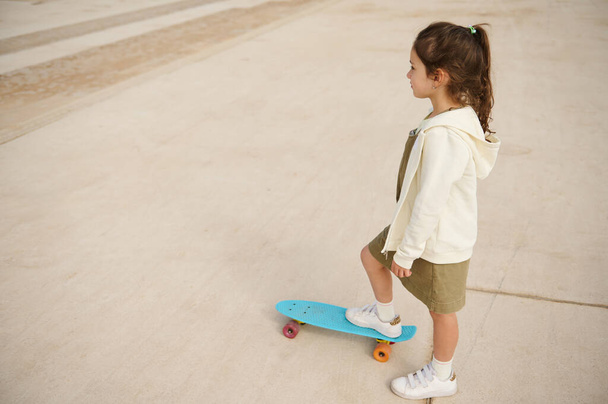 Вид сверху на кавказскую маленькую девочку, которая смотрит в сторону во время игры на скейтборде или катания на коньках и начинает на открытой насосной дорожке в скейтпарке экстремальными видами спорта. Людей. Детство. Отдых - Фото, изображение