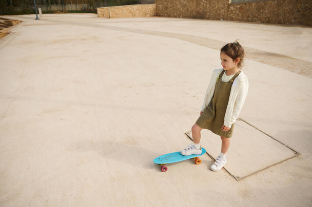 Vue d'en haut de la belle petite fille blanche debout sur son skateboard sur une jambe, regardant au loin, debout seul sur l'aire de jeux skatepark. Enfance. Loisirs - Photo, image
