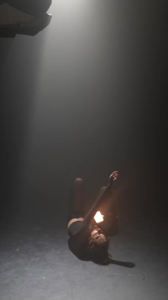 Dans une pièce faiblement éclairée, une jeune fille en tenue sombre danse activement, tenant une lumière dans ses mains. Images 4k de haute qualité - Séquence, vidéo