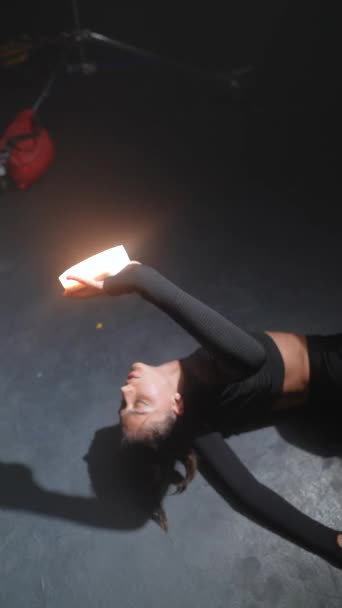 Avec une lumière à la main, une jeune femme en tenue sombre danse énergiquement dans un espace faiblement éclairé. Images 4k de haute qualité - Séquence, vidéo